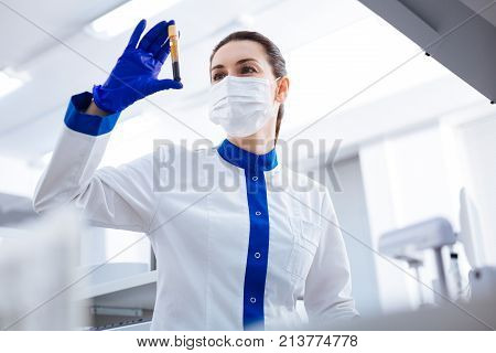 HIV test. Elegant professional female intern wearing mask while rising vial with blood and looking at it