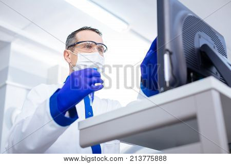 Data transferring. Confident handsome male scientist holding glassware putting data into the computer and looking at screen attentively