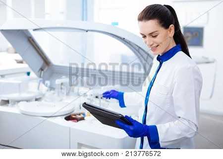 Science is light. Beautiful gentle inspired  female intern smiling adorably and looking at the tablet while stretching her hand to the centrifuge machine