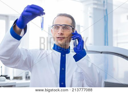 Phone conversation. Optimistic sweet male scientist talking on the phone while bringing up test tube with poured blue reagent
