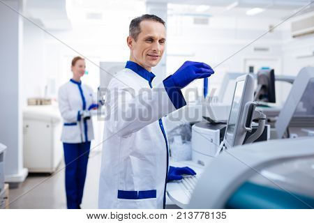 Let me see. Optimistic striking male researcher  smiling and typing while holding test glass in  a hand  near the medical equipment