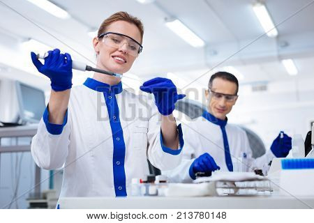 Routine work.  Elegant happy female researcher getting sample from the glassware carefully  while holding up pipette and standing in the laboratory
