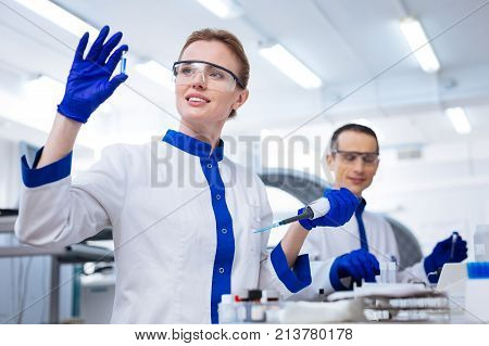 Biochemical lab. Glad inspired female lab assistant waiting for reaction after putting reagent into the sample with a pipette while wearing medical glasses