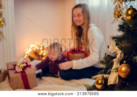 Christmas scene with happy family in blurred bokeh background. Teen sister with her baby brother in Christmas time open gifts and presents. Christmas tree on the foreground plan people out of focus.
