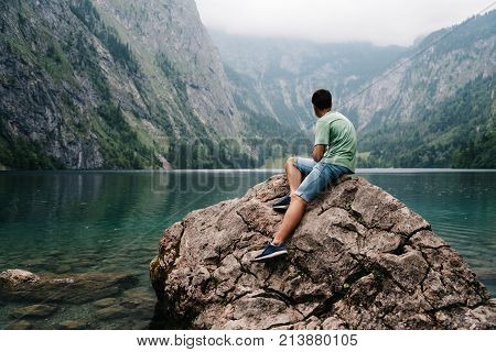 Young adult man sitting on rock looking at beautiful and misty lake in the alps. Obersee