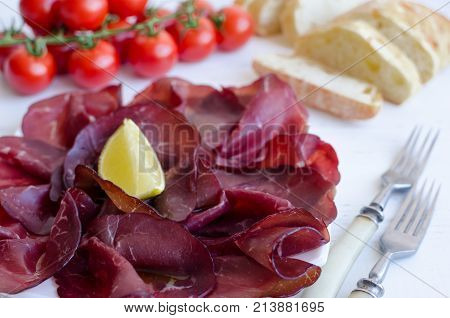 Slices of Italian meat Bresaola served with olive oil and lemon on a plate with tomatoes and bread on white background. Traditional appetizers antipasti. Top view.