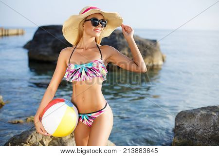 Young beautiful woman with a slim figure, blonde in dark sun glasses and a big straw light hat from the sun, motley bikini and a big inflatable ball, posing alone outdoors in the summer against a rocky beach and ocean