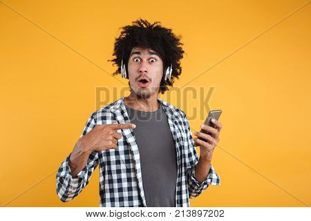 Portrait of an excited young african man listening to music with headphones while standing and poinitng finger at mobile phone isolated over orange background