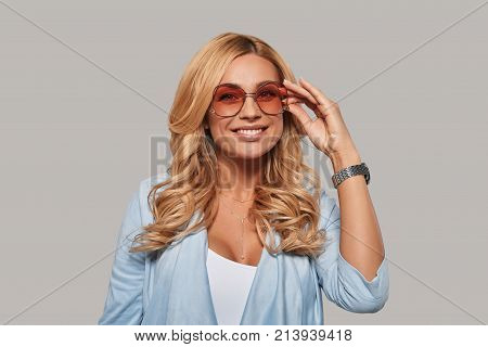 In her own style. Attractive young woman smiling and adjusting her eyewear while standing against grey background