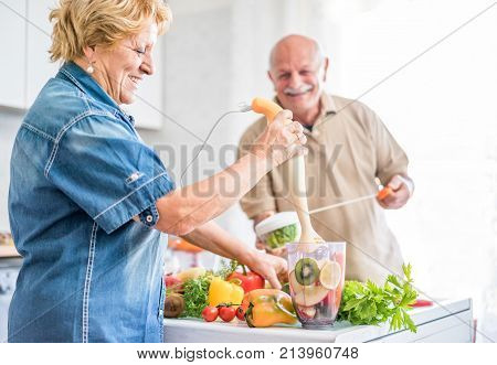Happy senior couple preparing healthy vegetarian breakfast with fruits and vegetables - Old cheerful people taking care about diet - Healthvegan and organic concept - Focus on woman face