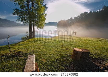 summer early morning on the lake in the fog. against the background of the mountains and the village on the shore. one girl shoots a landscape on a camera on the beach against a background of pink dawn