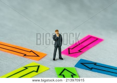 Miniature people figure businessman thinking and standing at the center of arrows pointing to him in all directions as business decision or people center concept.