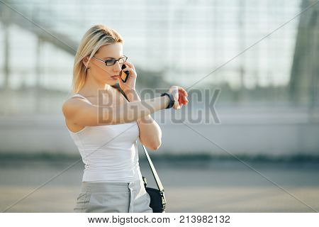 Attractive business woman using a phone and clock while standing in front of windows in an office building overlooking the city