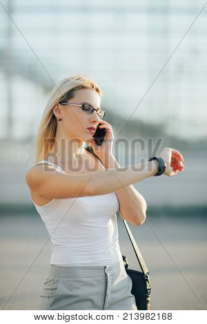 Attractive business woman using a phone and clock while standing in front of windows in an office building overlooking the city