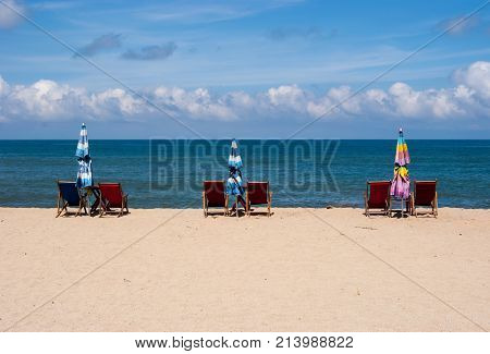 Beach lounge chairs in pairs with a folded up umbrella facing the ocean, beautiful lightly cloudy sky in the background