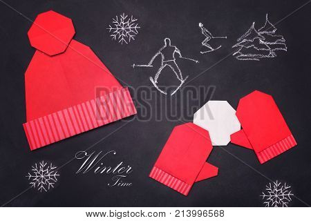 Origami red mitters and cap of paper on black background with skier and snowflake winter