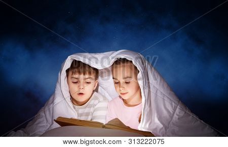 Engrossed Little Girl And Boy Reading Book In Bed After Bedtime. Pretty Kids Together Hiding Under B
