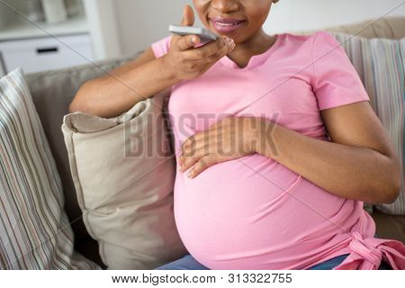pregnancy, technology and people concept - happy pregnant african american woman using voice command recorder or calling on smartphone at home