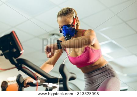 Low angle view of beautiful young Caucasian female athlete with oxygen mask checking time while exercising with exercise bike in fitness studio. Bright modern gym with fit healthy people working out