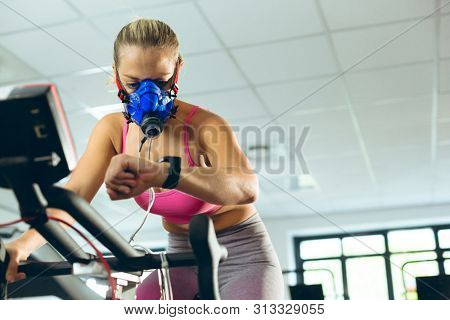 Low angle view of beautiful young Caucasian female athlete with oxygen mask checking time while exercising with exercise bike in fitness studio. Bright modern gym with fit healthy people working out