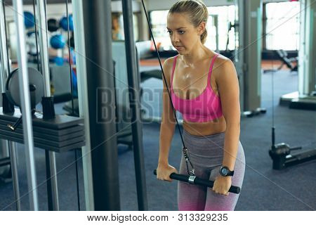 Front view of beautiful young Caucasian female athlete exercising with triceps machine in fitness studio. Bright modern gym with fit healthy people working out and training