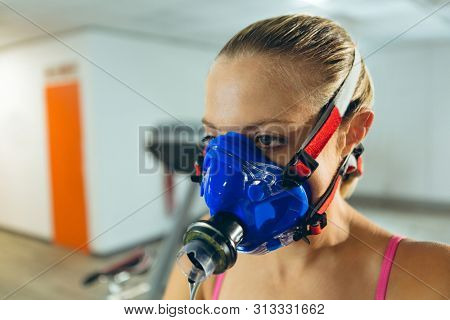 Close-up of beautiful young Caucasian woman with oxygen mask before exercise in fitness center. Bright modern gym with fit healthy people working out and training
