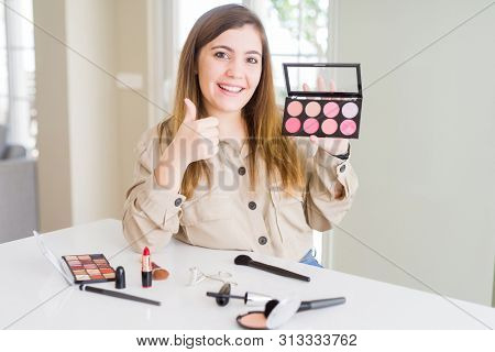 Beautiful young woman using make up cosmetics applying color from palete happy with big smile doing ok sign, thumb up with fingers, excellent sign