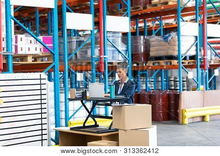 Front view of beautiful mature Caucasian female manager working at desk in workhouse. This is a freight transportation and distribution warehouse. Industrial and industrial workers concept