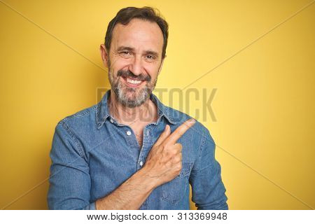 Handsome middle age senior man with grey hair over isolated yellow background cheerful with a smile of face pointing with hand and finger up to the side with happy and natural expression on face