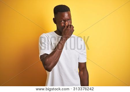 Young african american man wearing white t-shirt standing over isolated yellow background smelling something stinky and disgusting, intolerable smell, holding breath with fingers on nose. Bad smells