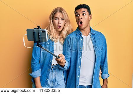 Young interracial couple taking a selfie photo with smartphone afraid and shocked with surprise and amazed expression, fear and excited face. 
