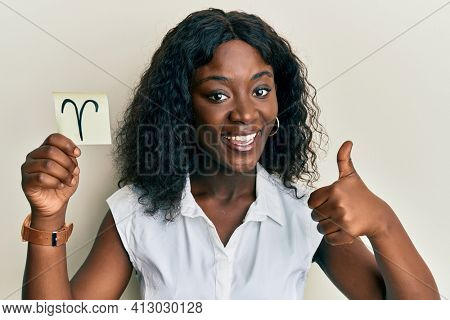 Beautiful african young woman holding paper with aries zodiac sign smiling happy and positive, thumb up doing excellent and approval sign 