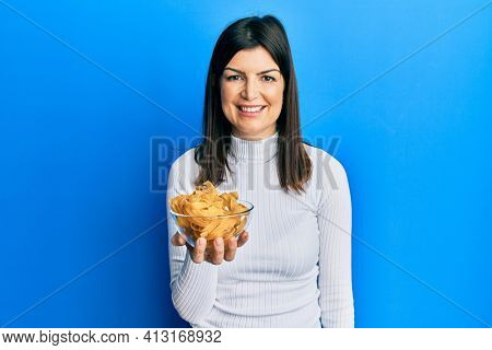 Young hispanic woman holding uncooked pasta bowl looking positive and happy standing and smiling with a confident smile showing teeth 