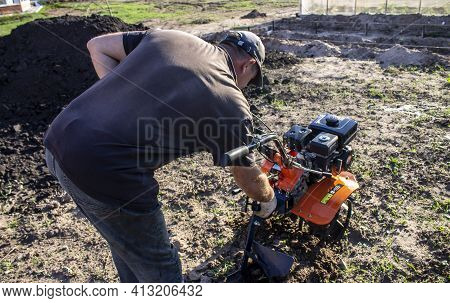 Male In The Spring Plows The Earth In Working Clothes With A Motoblock, Walk-behind Tractor