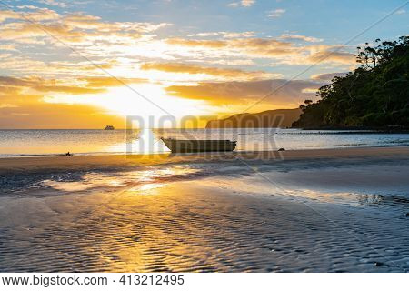 Setting Sun Glistens On Water And Sand Patterns With Dinghy Silhouette On Beach At Low Tide On Mulbe