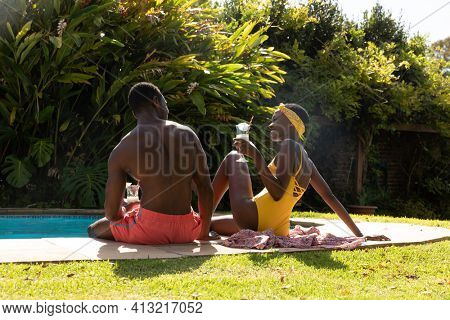 African american couple sitting at the poolside and drinking drinks on sunny garden terrace. staying at home in isolation during quarantine lockdown.