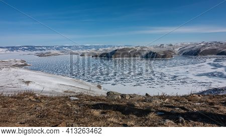 The Frozen Lake Baikal Is Covered With Lace-like Snow. In The Foreground Is The Earth, Dry Grass. In