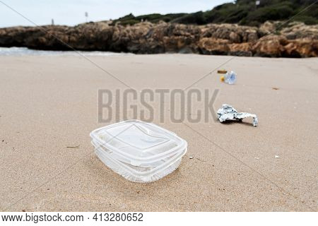 closeup of a used plastic container thrown on the wet sand of a beach, next to some other garbage, such as a piece of aluminum foil or a used plastic bottle