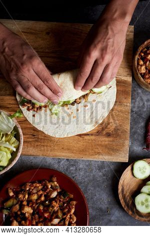 a young caucasian man, seen from above, rolls a durum or a burrito, filled with chicken meat cooked with different vegetables such as onion or red and green pepper, and fresh lettuce and raw cucumber