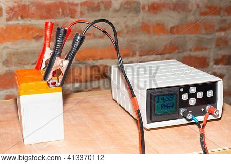 Charging The Motorcycle Battery. On A Wooden Surface Against The Background Of A Brick Wall Of The G