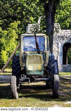 Old Tractor With Big Wheels And Rusty Exhaust Pipe. It Stands On Background Of Green Trees. Close-up