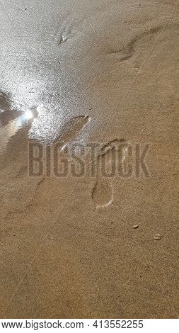 Footprints Of Human Feet On The Sand Near The Water On The Sea Beach.