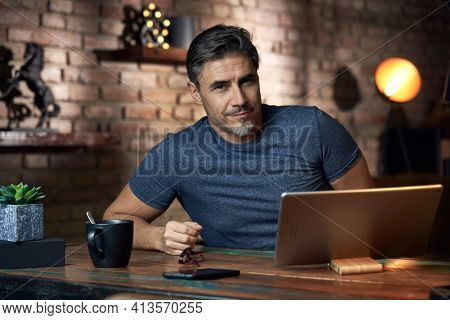 50s man working online with laptop computer at home sitting at desk, smiling. Home office, browsing internet, study room, Businessman working from home.