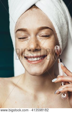 Freckled Woman Is Using A Facial Massage Roller Smiling While Covering Head With Towel And Posing Wi