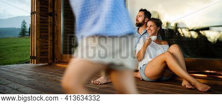 Family With Small Daughter Sitting On Patio Of Wooden Cabin, Holiday In Nature Concept.