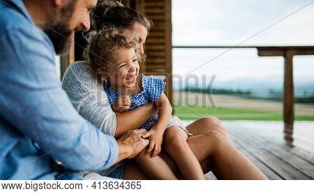 Family With Small Daughter Sitting On Patio Of Wooden Cabin, Holiday In Nature Concept.