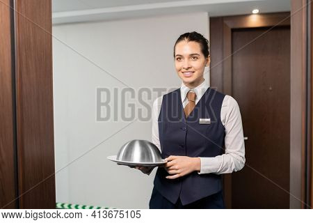 Happy young elegant manager in uniform holding tray with breakfast while standing in front of open door in the corridor of luxurious hotel