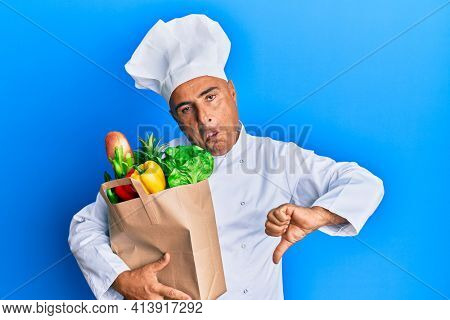 Mature middle east man professional chef holding bag of groceries with angry face, negative sign showing dislike with thumbs down, rejection concept 