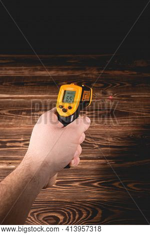 A Man Holds A Yellow-black Pyrometer On A Wooden Background. A Device For Non-contact Temperature Me