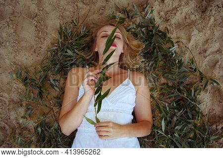 Top View Of A Woman In A White Dress With A Long Branch With Green Leaves On Her Face Lies On The Sa
